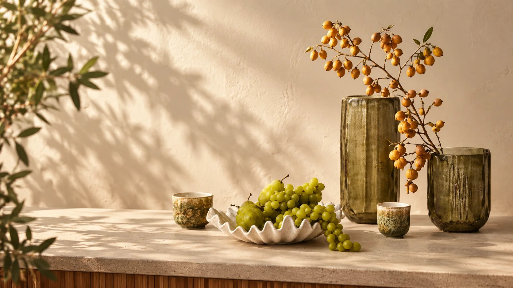 Olive glass vessels with orange berry stems and a shell bowl of fruit on a stone console